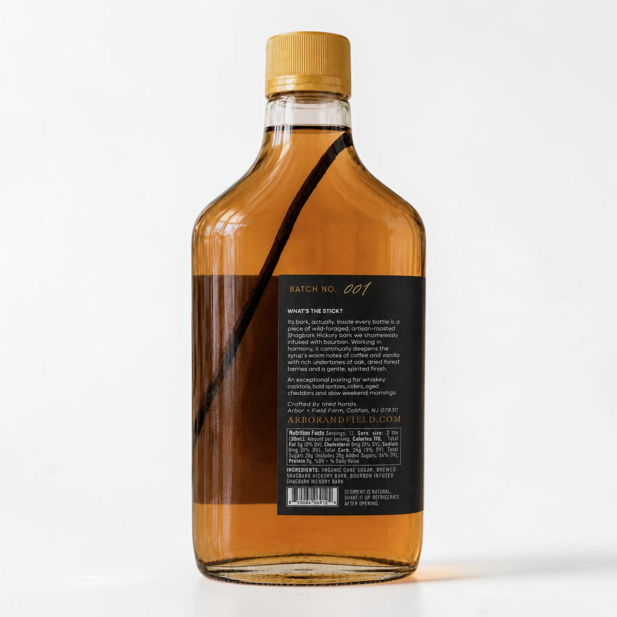 Bottle of Arbor and Field Black Label Bourbon-Infused Shagbark Hickory Syrup with black and gold foil label. A piece if bourbon-infused shagbark hickory bark can be seen inside of the bottle. The image shows the back label of the bottle with a handwritten gold batch number, pairing suggestions and a description of how the product is crafted. Clean white background, premium look and feel.