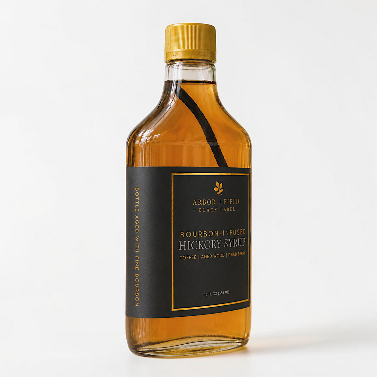 Bottle of Arbor and Field Black Label Bourbon-Infused Shagbark Hickory Syrup with black and gold foil label. A piece if bourbon-infused shagbark hickory bark can be seen inside of the bottle. The spine of the bottle reads "bottle aged with fine bourbon". Clean white background, premium look and feel.