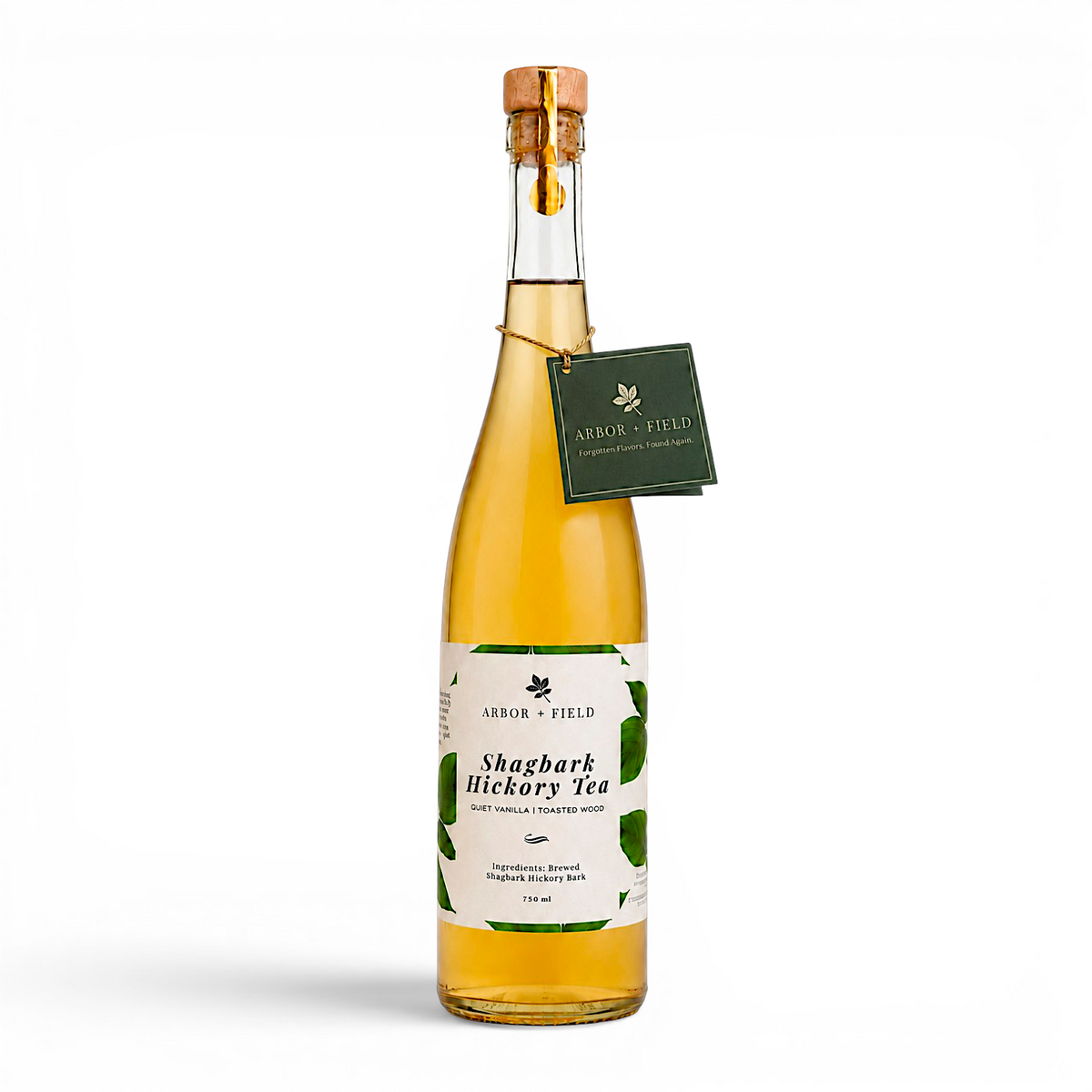 Bottle of Arbor and Field shagbark hickory tea with a wooden cork, green and white label and a green gift tag hanging from the neck by a gold string. Modern clear glass wine bottle with amber colored tea inside. Clean white background, premium look and feel.