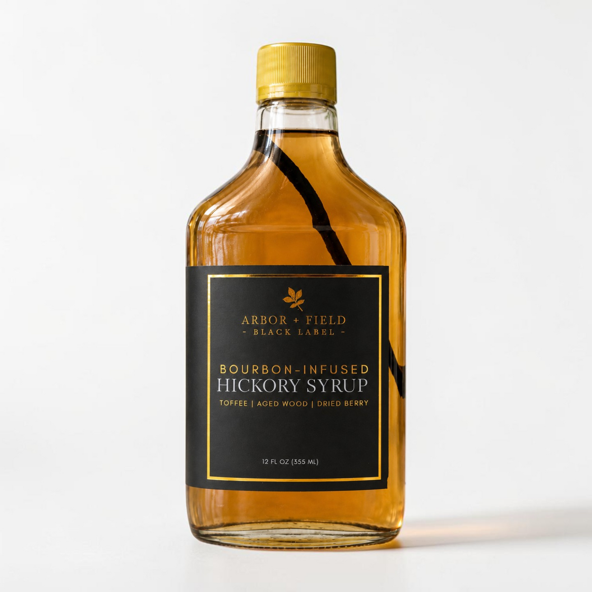 Bottle of Arbor and Field Black Label Bourbon-Infused Shagbark Hickory Syrup with black and gold foil label. A piece if bourbon-infused shagbark hickory bark can be seen inside of the bottle. Clean white background, premium look and feel.
